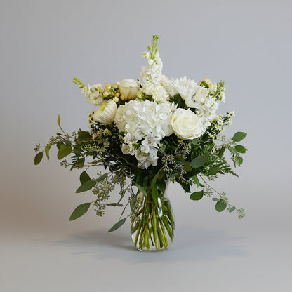 A white and green floral arrangement in a clear glass vase, featuring white roses, hydrangeas, and eucalyptus leaves against a plain light background.